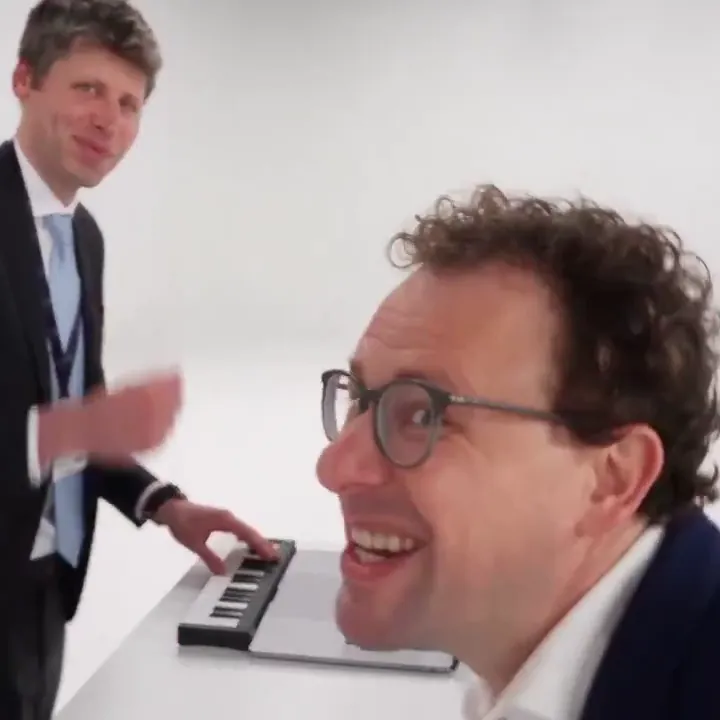 A smiling man in glasses leans into the lens so his face fills the frame, with his friend still at the keyboard behind him