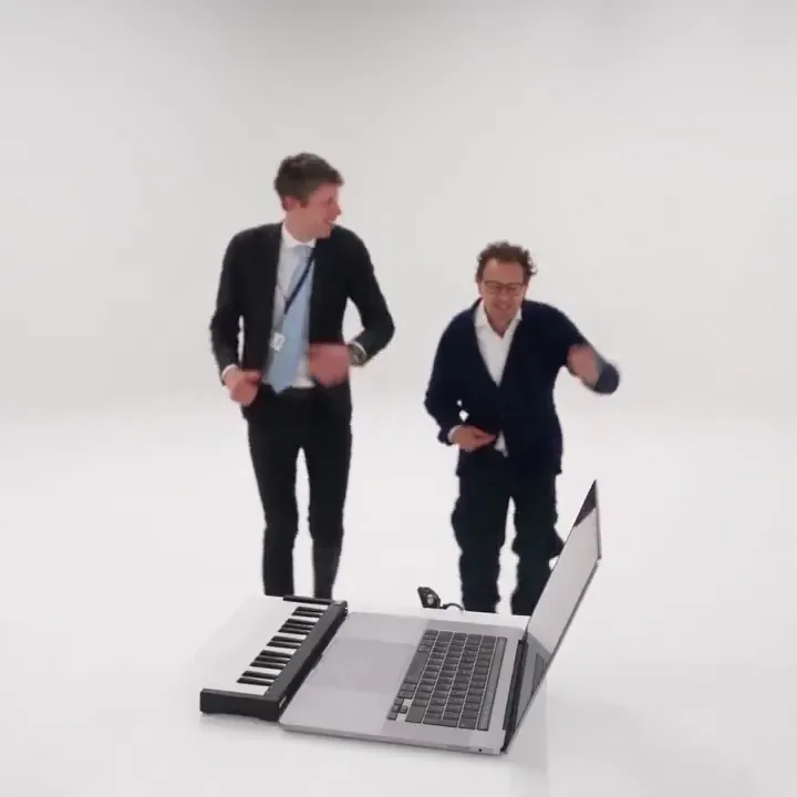 Two men in suits dance away from the laptop and mini keyboard across a white studio