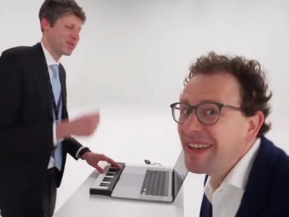A man in glasses grins straight into the camera while his friend plays the mini keyboard behind him