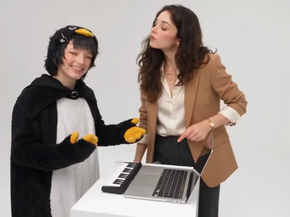 A grinning person in a penguin onesie waves the costume's yellow flippers at the keyboard while a friend in a tan blazer looks on