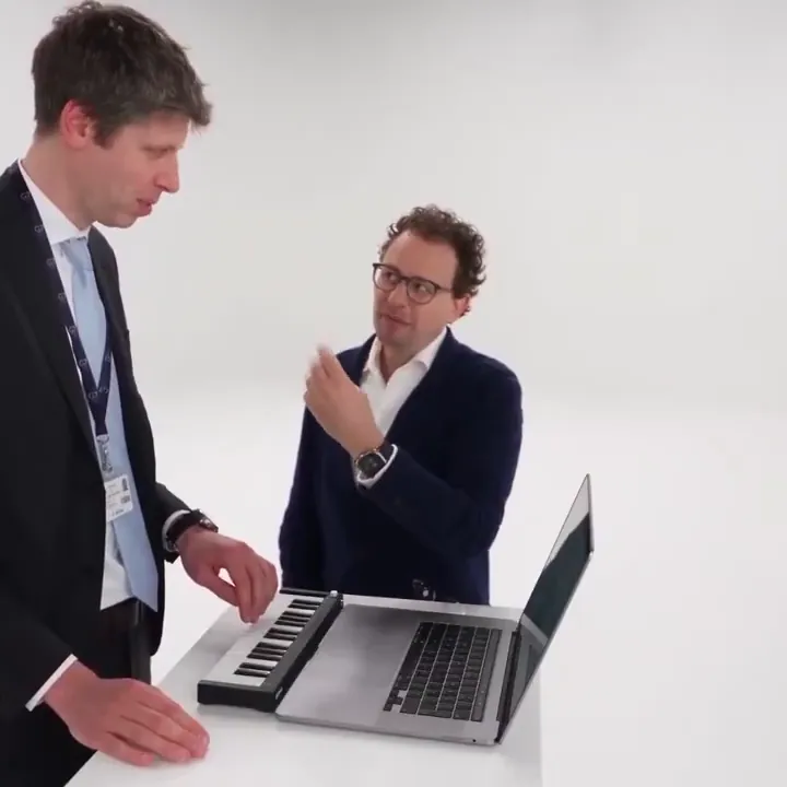 Two men at a laptop with a mini keyboard in a white studio, one playing while the other gestures
